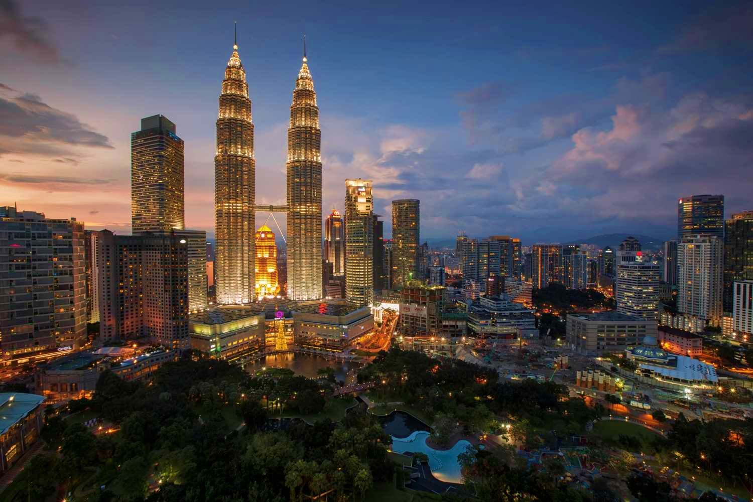 Kuala Lumpur skyline at dusk with illuminated Petronas Twin Towers, surrounding skyscrapers, and Klcc park with blue pool in 
